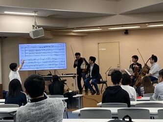 ステージ上で、左からフルート、ヴァイオリン、ヴィオラ、チェロの演奏家が並び、演奏曲の楽譜について説明している様子