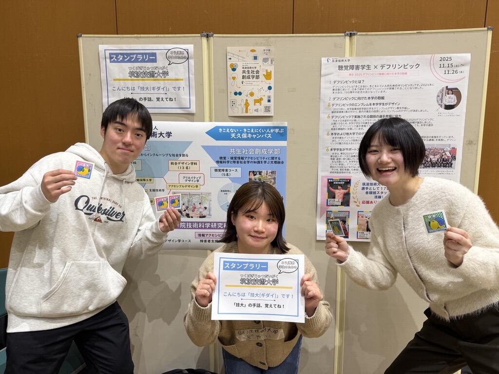 本学を紹介するパネルの前で記念撮影をしたブース担当の学生3名